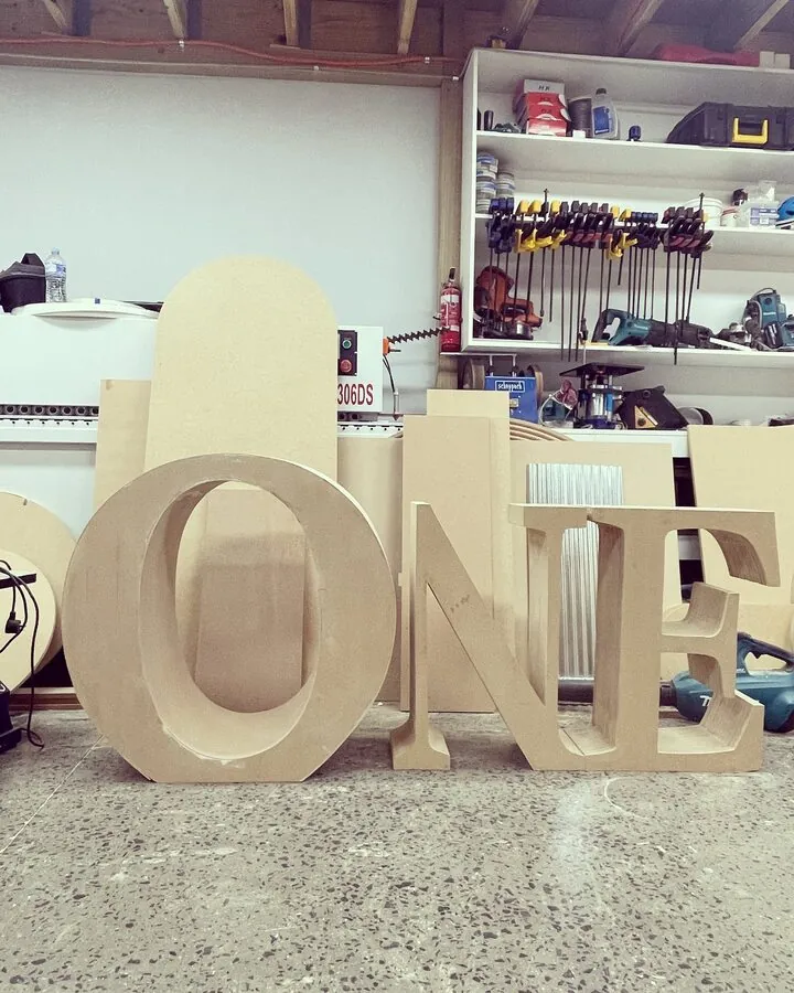 Giant MDF letters spelling ONE, freshly cut and standing in the workshop